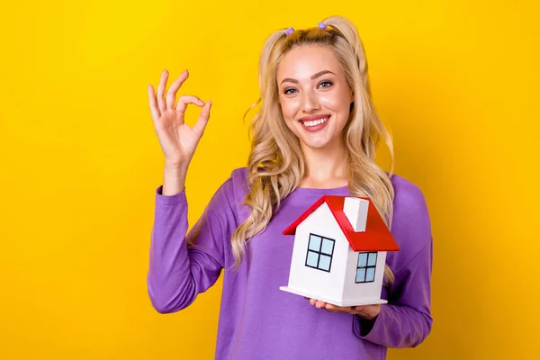 迷人而自信的女人穿着紫色衬衫 拿着一幢小房子 摆出一副奇怪的姿势 黄褐色背景的照片 — 图库照片