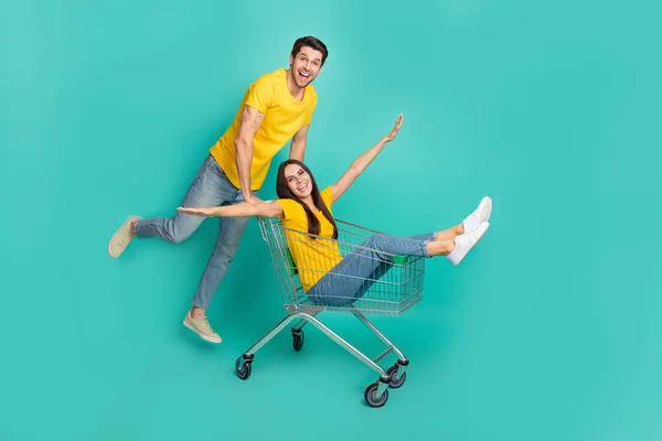 かなり屈託のない夫婦の写真は楽しい孤立したティール色の背景を持つショッピングトレイに乗って黄色のTシャツを身に着けています — ストック写真