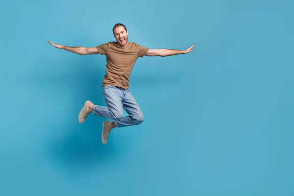 Full length photo of pretty impressed guy dressed beige t-shirt jumping high arms sides empty space isolated blue color background.