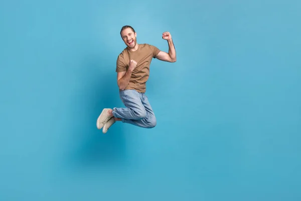 Full length photo of cute lucky guy dressed beige t-shirt jumping high rising fists empty space isolated blue color background.