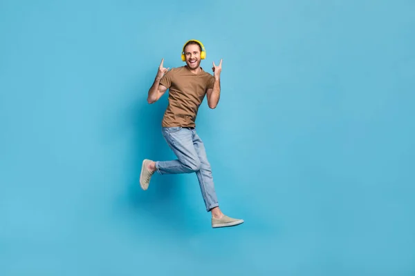 Full length photo of funky cool guy dressed beige t-shirt jumping listening hard rock gesture isolated blue color background.