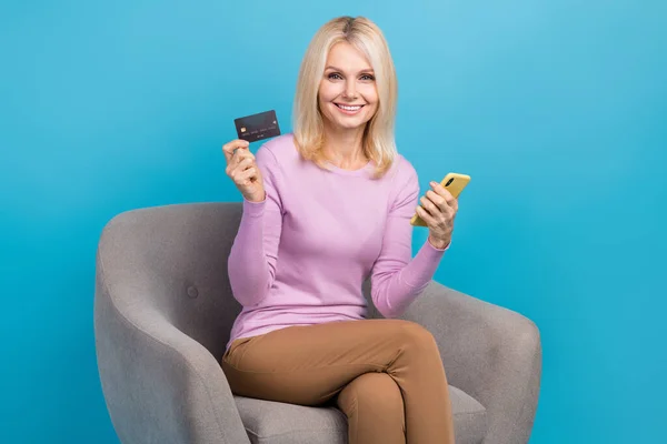 Photo of adorable positive lovely lady sitting office chair hold bank debit card recommend easy payment isolated on blue color background.