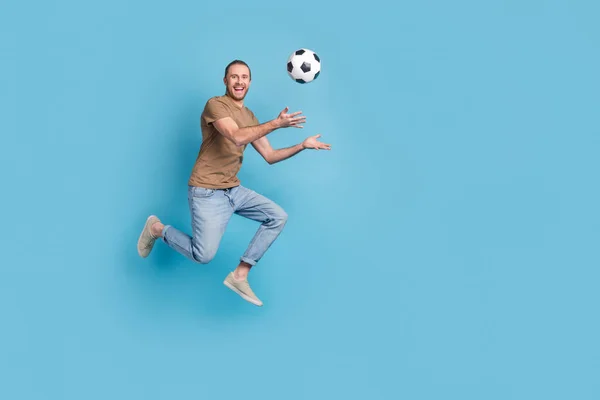 Full length photo of positive excited man wear beige t-shirt jumping high playing football empty space isolated blue color background.