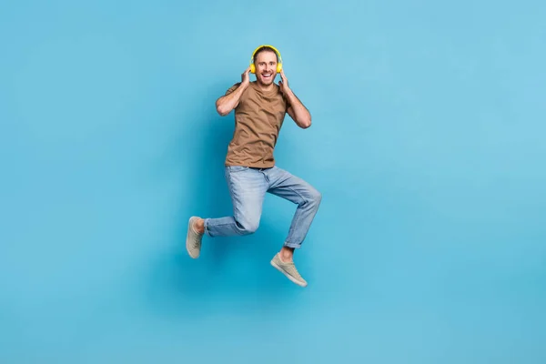 Full length photo of excited cheerful man wear beige t-shirt jumping high enjoying music empty space isolated blue color background.