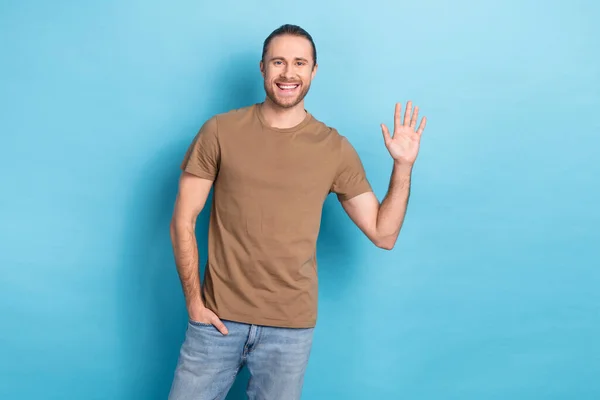 Portrait of satisfied glad person toothy smile put hand pocket palm waving isolated on blue color background.