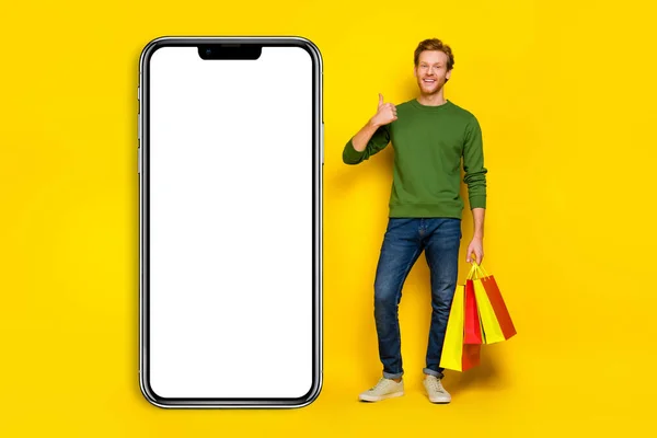 Full length photo of excited cool man wear pullover bargains thumb up gadget empty space isolated yellow color background.