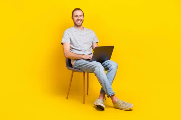 楽観的なスマート男の完全な長さの写真は ライトシャツのジーンズスニーカーは黄色の色の背景に隔離された椅子に座ってラップトップを保持 — ストック写真