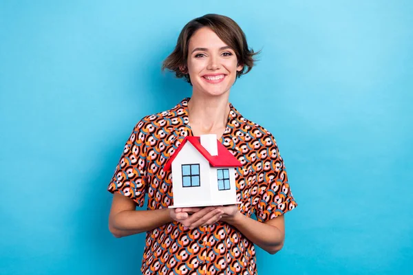 Photo of pretty cheerful woman wear print blouse smiling holding small red roof house isolated blue color background.