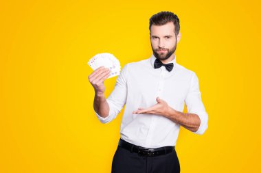 Portrait of stylish trendy croupier with hairstyle in formal wear having, showing with palm set of cards in hands, isolated on grey background, looking at camera. clipart