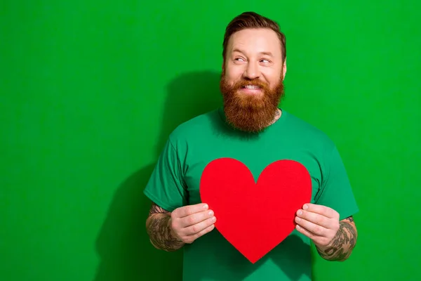Portrait of cheerful satisfied man with red beard wear trendy t-shirt hold red heart look empty space isolated on green color background.