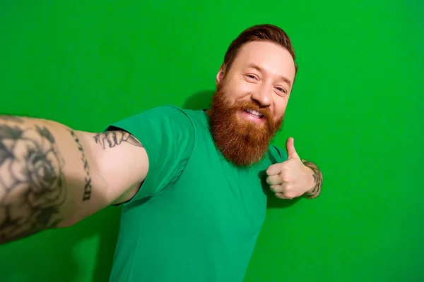 Photo of nice handsome guy with ginger beard stylish t-shirt doing selfie showing like good job isolated on green color background.