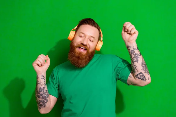 Photo of good mood nice guy with ginger beard dressed stylish t-shirt headphones listening playlist isolated on green color background.