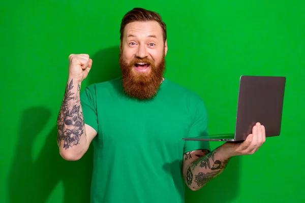 赤いひげを生やした残忍な満足男の肖像は流行のTシャツを保持ノートパソコンの叫びはい勝つ賭け緑の色の背景に隔離された — ストック写真