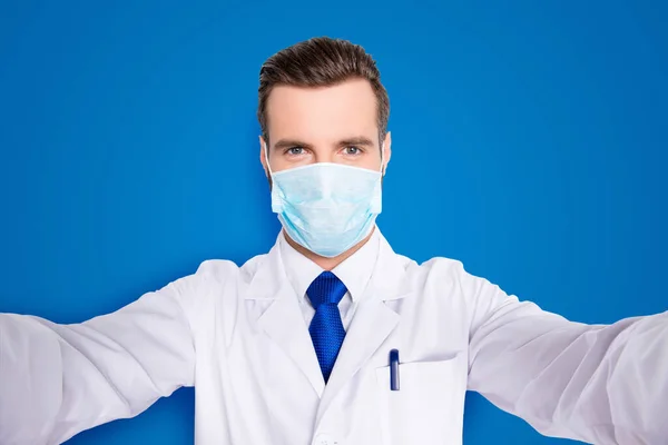 Self portrait of creative funny dentist in white lab coat, blue, tie, protective face mask shooting selfie on smart phone, having rest relax video call, isolated on grey background.
