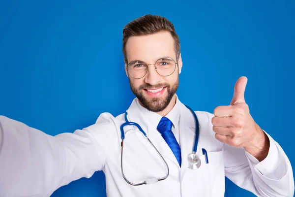 Self portrait of positive trendy doc in white outfit with tie and bristle having stethoscope on his neck shooting selfie showing thumb up, like sign with finger, hand over grey background.