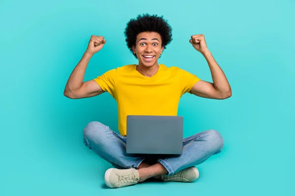 Full body photo of delighted excited person sit floor raise fists use netbook isolated on turquoise color background.