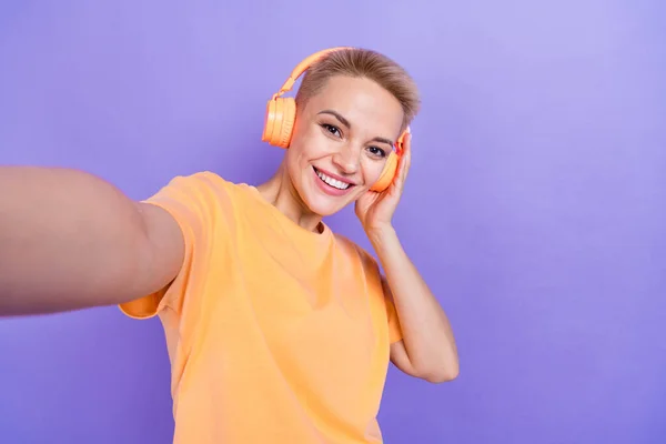 stock image Portrait of cheerful woman with blond hairstyle wear orange t-shirt make selfie hand touch headphones isolated on violet color background.