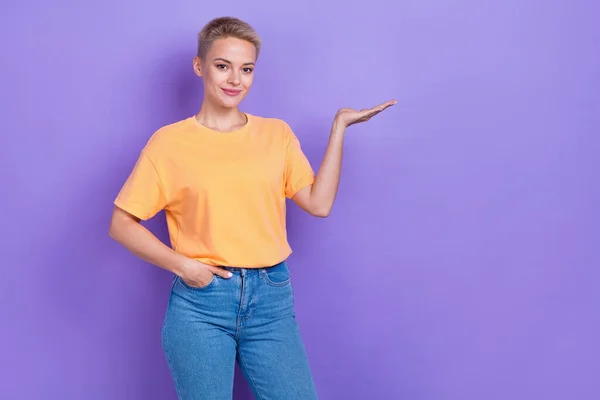 真剣なプロモーターの写真若い女性はオレンジのTシャツデニムジーンズを着用アーム製品ブランド名モックアップは 紫色の背景に隔離された保持しています — ストック写真
