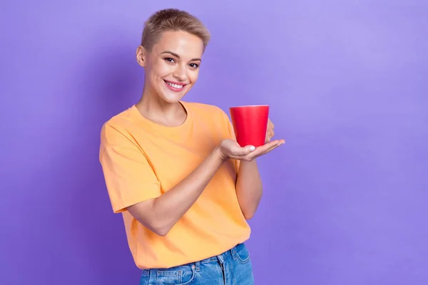 ブロンドの髪型と良い気分の女性の肖像は 紫の色の背景に隔離された朝のコーヒーのオレンジTシャツホールドカップを身に着けている — ストック写真