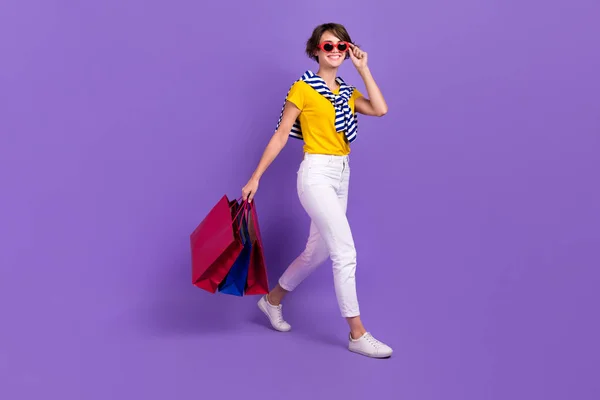 Full length photo of positive pretty lady wear bright clothes go empty space carry packages packs isolated on purple color background.