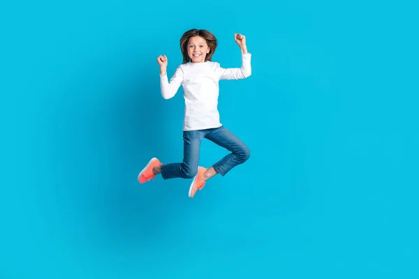 Full size photo of overjoyed ecstatic girl wear white long sleeve denim trousers jump celebrate winning isolated on blue color background.