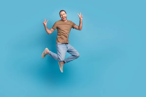 Full length photo of excited funny man wear beige t-shirt jumping high showing v-signs emtpy space isolated blue color background.