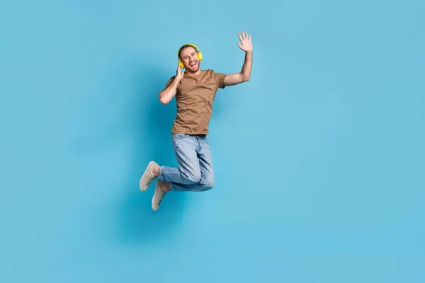 Full length photo of cheerful dreamy man wear beige t-shirt jumping high listening songs empty space isolated blue color background.
