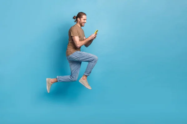 Full length photo of excited pretty guy dressed t-shirt jumping typing apple samsung gadget empty space isolated blue color background.