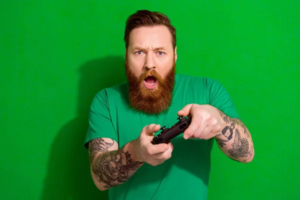 Portrait of masculine man red beard wear trendy t-shirt holding playstation joystick play excited game isolated on green color background.