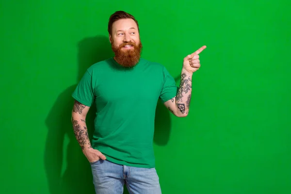 Photo of positive guy with ginger beard dressed stylish t-shirt indicating look billboard empty space isolated on green color background.