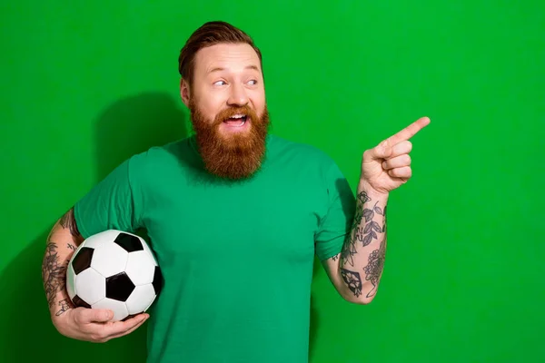 Photo of impressed guy ginger beard dressed stylish t-shirt hold soccer ball look directing empty space isolated on green color background.