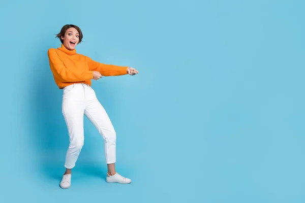 stock image Full size photo of pretty young girl pulling rope string excited empty space dressed stylish orange look isolated on blue color background.