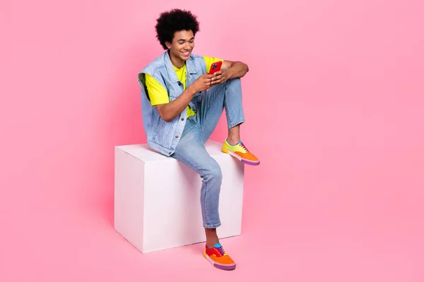 Full length photo of positive man blogger wear bright clothes sitting podium use telephone empty space isolated on pink color background.