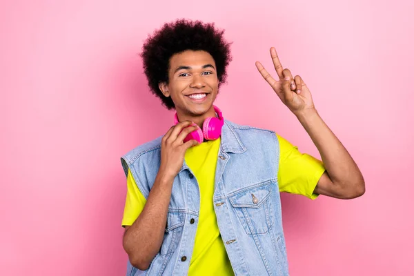 Portrait of cheerful friendly person toothy smile show v-sign touch headphones hang neck isolated on pink color background.