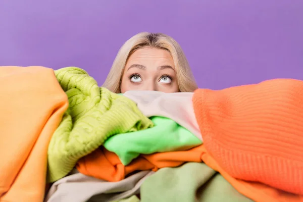 Photo of young lady hide face behind big pile stack clothes look up empty space isolated on violet color background.