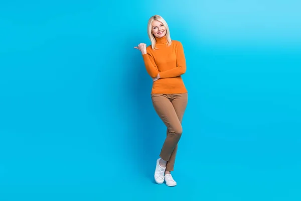 満足した女性のフルサイズの写真は 青の色の背景に隔離された販売空のスペースで示すオレンジ色のタートルネック茶色のズボンを身に着けている — ストック写真