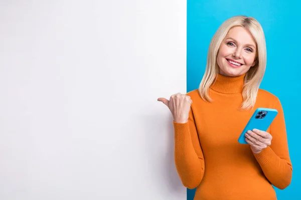 Photo of pensioner attractive blonde woman wear orange turtleneck direct finger placard customer ad isolated on blue color background.