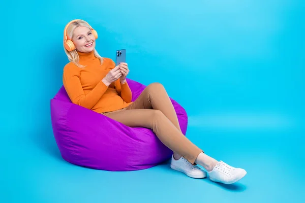 楽観的な女性の完全な長さの写真オレンジ色のスウェットシャツはヘッドフォンでPoufに座っている青い色の背景に隔離されたスマートフォンを保持 — ストック写真