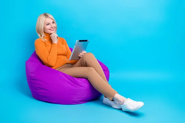 Full size photo of intelligent woman orange turtleneck sitting on pouf hold tablet look empty space isolated on blue color background.