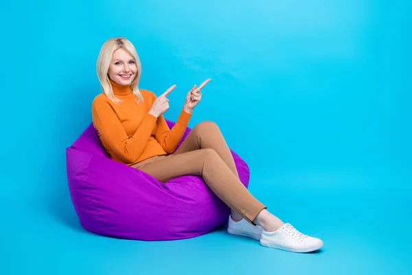 フルサイズの写真の面白い女性オレンジTurtleeck座っています上のPouf示すで提供の空のスペース隔離上の青の色の背景 — ストック写真