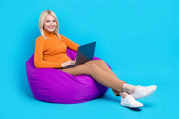 Full size photo of peasant woman dressed orange turtleneck sitting on pouf hold laptop on interview isolated on blue color background.