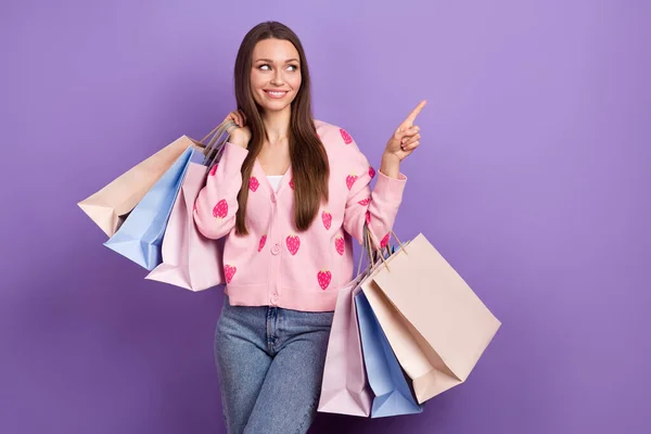 购物狂的年轻女子沉迷于购买更多衣服的照片直接用手指看空旷的空间拿着用紫色背景隔开的包 — 图库照片