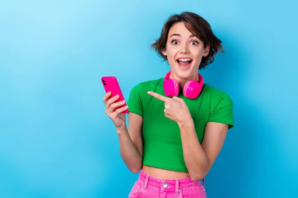 stock image Photo of funny youngster girl wear green stylish t-shirt direct finger her itunes listen music bluetooth earphones isolated on blue color background.