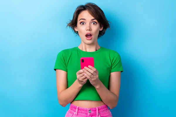 年轻而震惊的美女穿着绿色T恤粉红裤子拿着智能手机震惊的打折商店蓝色背景的照片 — 图库照片