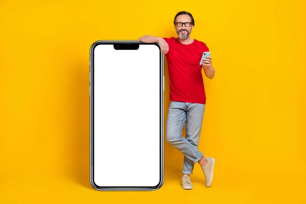 Photo of charming pretty guy pensioner dressed red t-shirt glasses typing gadget empty space isolated yellow color background.