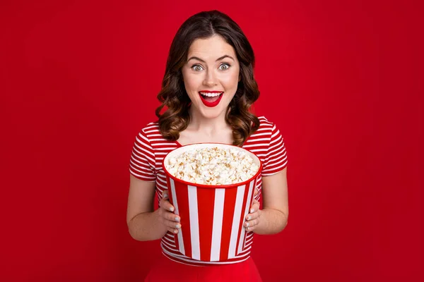 Photo of overjoyed funny lady watch new bestseller movie hold big popcorn bucket wear striped top isolated red color background.