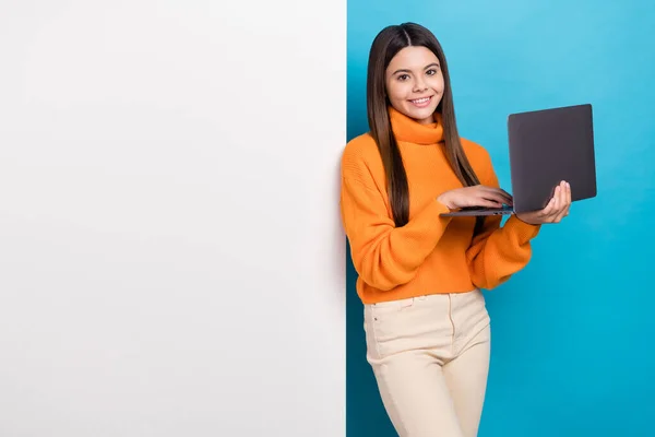 满意的女孩直着头发 穿着橙色毛衣 手持笔记本电脑 站在空旷的空间广告牌旁边 蓝色背景隔离 — 图库照片