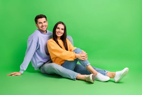 甜蜜的年轻夫妇穿着运动衫坐在地板上 背负着孤零零的绿色背景的全长照片 — 图库照片