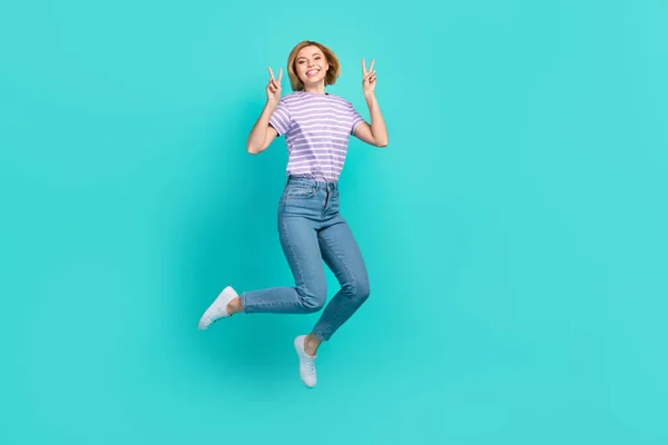 Full length photo of gorgeous girl dressed stylish t-shirt denim pants jumping showing v-sign isolated on turquoise color background.