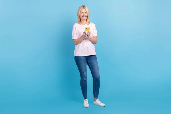 明るい人の完全な長さの写真を身に着けている白いTシャツデニムパンツは 青の色の背景に隔離されたスマートフォンで通知を読む — ストック写真
